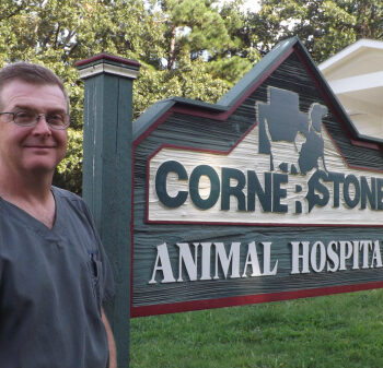 Dr. Marty Turner smiling while standing outdoors next to the large wooden Cornerstone Animal Hospital sign, with the clinic building and trees in the background – Cornerstone Animal Hospital